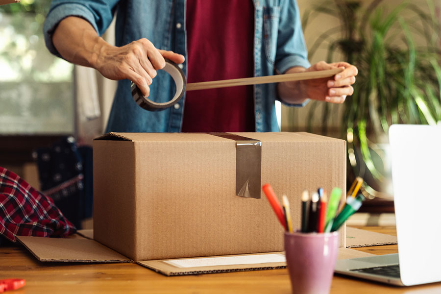 Person sealing a cardboard box with tape at a desk, surrounded by packing items